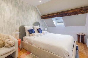 Ein Schlafzimmer mit schrägen Wänden, einem Bett mit weißer Bettwäsche, einem grauen Sofa, einem kleinen runden Tisch und einer Wand mit Muster.