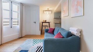 A modern living room with a sofa, a dining area, and a kitchen in the background. - 11 Beaumarchais 5D (Paris)