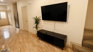 Een woonkamer met een flatscreen televisie aan de muur, een zwart tv-meubel, een potplant en een deur naar een andere kamer.