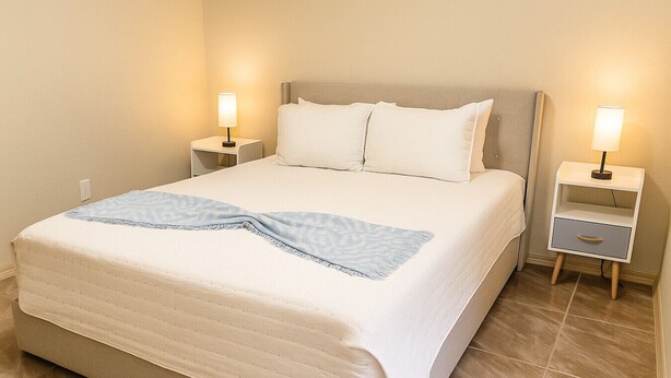 Een net opgemaakt bed met een blauwe plaid, twee nachtkastjes met lampen, en een klein dressoir.