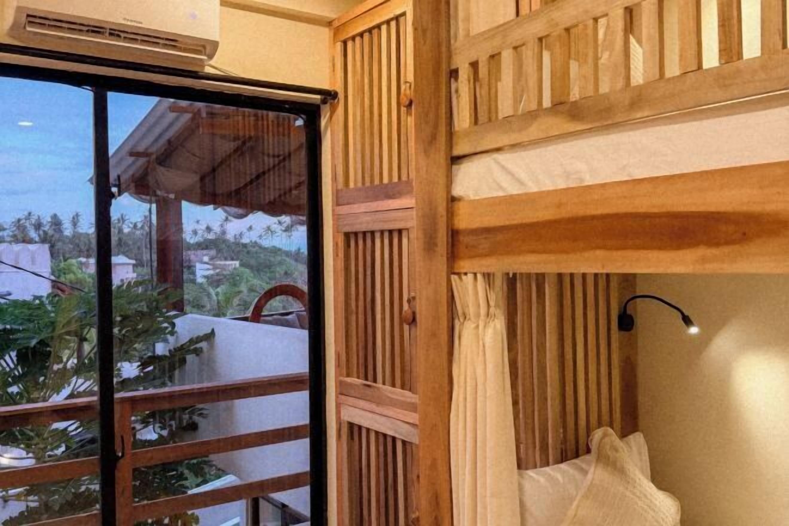 A room with a wooden slatted wall, a window with a view of greenery, and a bed with a white pillow.