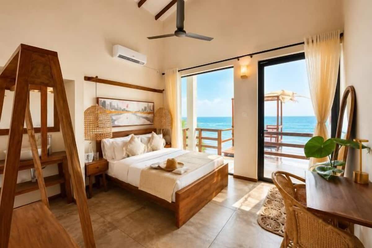 A bedroom with a wooden bed, a ceiling fan, a large window with a view of the sea, and a balcony with a chair.