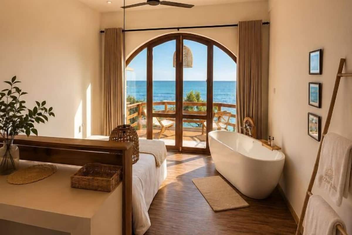 A bedroom with a large window, a bathtub, and a wooden deck with outdoor furniture.