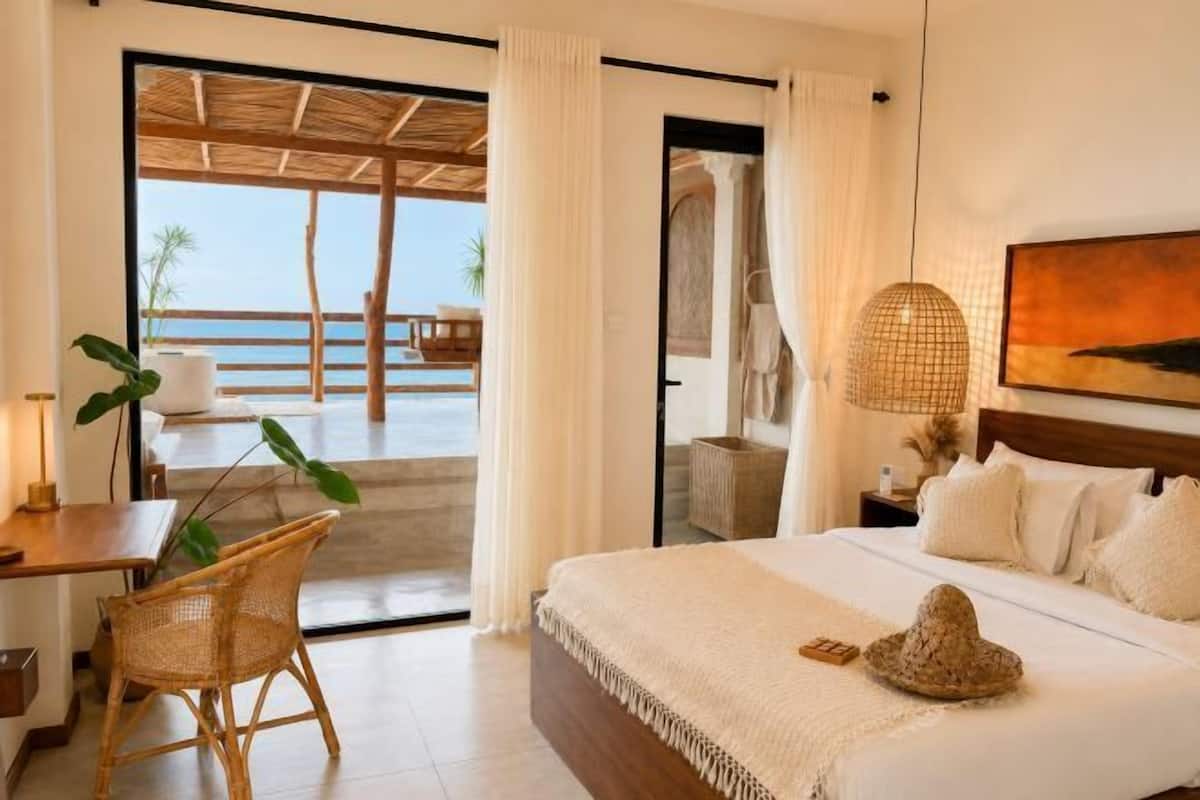 A bedroom with a bed, a desk, a chair, a large window, and a view of the beach.