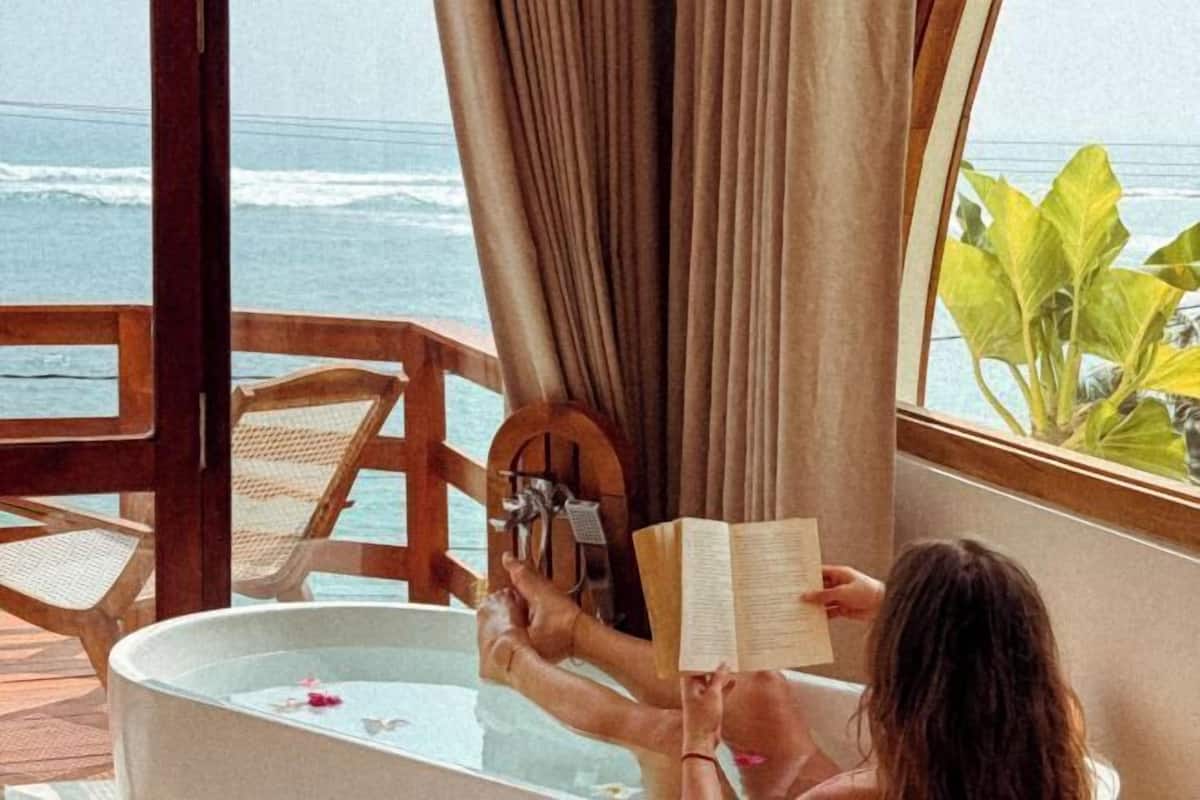 A person reading in a bathtub with a view of the ocean.