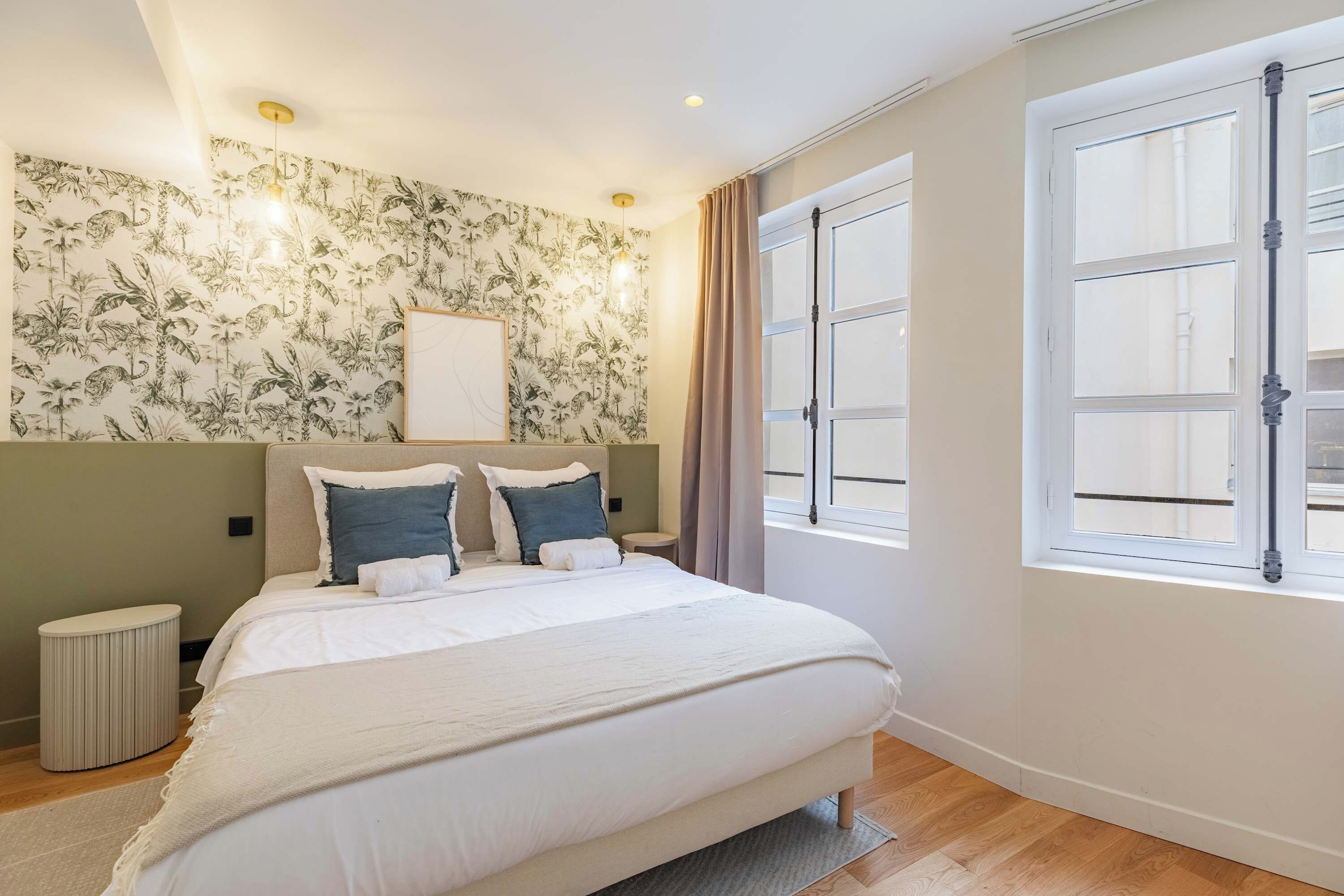 Une chambre à coucher comprenant un lit, une table de chevet, une lampe fixée au mur, une fenêtre avec des rideaux et un mur recouvert d’un papier peint à motifs tropicaux.