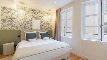 Une chambre à coucher comprenant un lit, une table de chevet, une lampe fixée au mur, une fenêtre avec des rideaux et un mur recouvert d’un papier peint à motifs tropicaux.