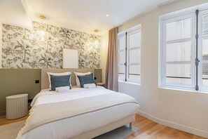 Un dormitorio con cama, mesita de noche, luz de pared, ventana con cortinas y papel tapiz tropical.