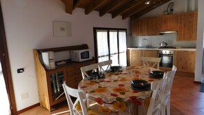 A dining area with a table set for four, surrounded by white chairs. A kitchen with wooden cabinets and a stove is visible in the background. - Rosemary 2 apartment in Agra with pool (Agra)