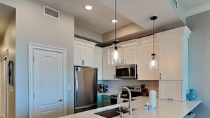 Cozinha moderna com armários brancos, eletrodomésticos de aço inoxidável, uma ilha central com banquetas, iluminação pendente e micro-ondas sobre a pia.