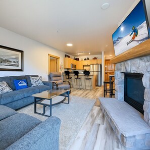 A modern living room with a sofa, coffee table, and fireplace. A kitchen with wooden cabinets and a stone fireplace is visible in the background. - Mountain Escape  | Walk to Village & Summer Fun (Keystone)