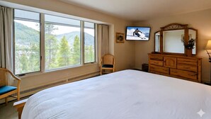 Habitación de hotel con una cama grande, televisión, cómoda, silla y ventana con vista a la montaña.