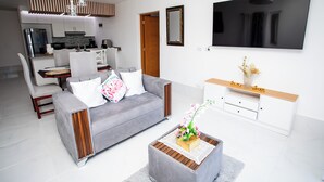Una sala de estar moderna con un sofá gris, una mesa de centro con un florero, un televisor montado en la pared y una cocina al fondo.