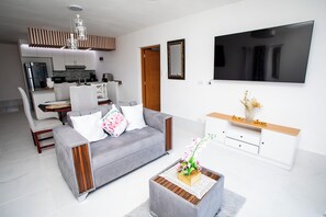 A modern living room with a grey sofa, a coffee table with a vase, a TV mounted on the wall, and a kitchen in the background. - Apartment with pool and view, minutes away from Cofresí and Costambar beaches. (Cofresi)