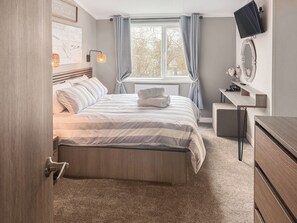 A bedroom with a bed, a desk, a television, and a window with curtains. - 38 at the Sanctuary Lodges - UK50850 (Rhosfawr, near Pwllheli)