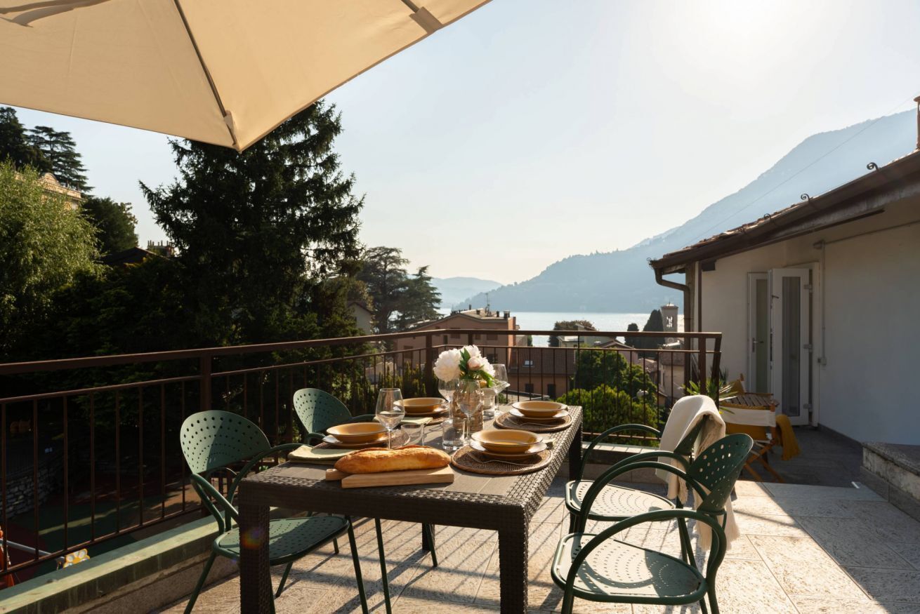 Un espace de restauration en plein air avec une table dressée pour deux personnes, entourée de chaises, d’un grand parasol et offrant une vue magnifique sur un lac et des montagnes.