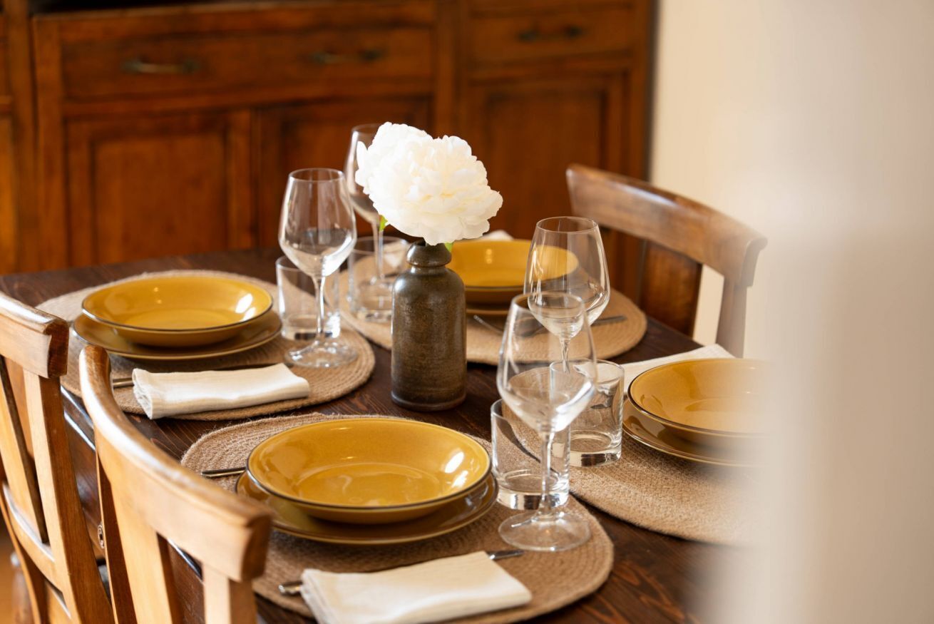 A dining table set with yellow plates, wine glasses, and a vase with a white flower.