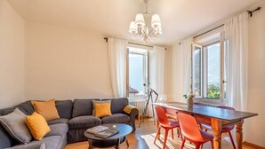 Een woonkamer met een bank, een eettafel met stoelen, een klein rond tafeltje en een kroonluchter.