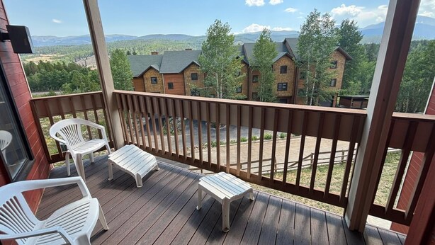Una terraza de madera con dos sillas de plástico blancas y una mesita, con vista a una hilera de casas de ladrillo con techos verdes y un paisaje montañoso.