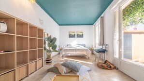 Une chambre à coucher avec un lit, une étagère en bois, une plante en pot et une fenêtre donnant sur un paysage verdoyant.