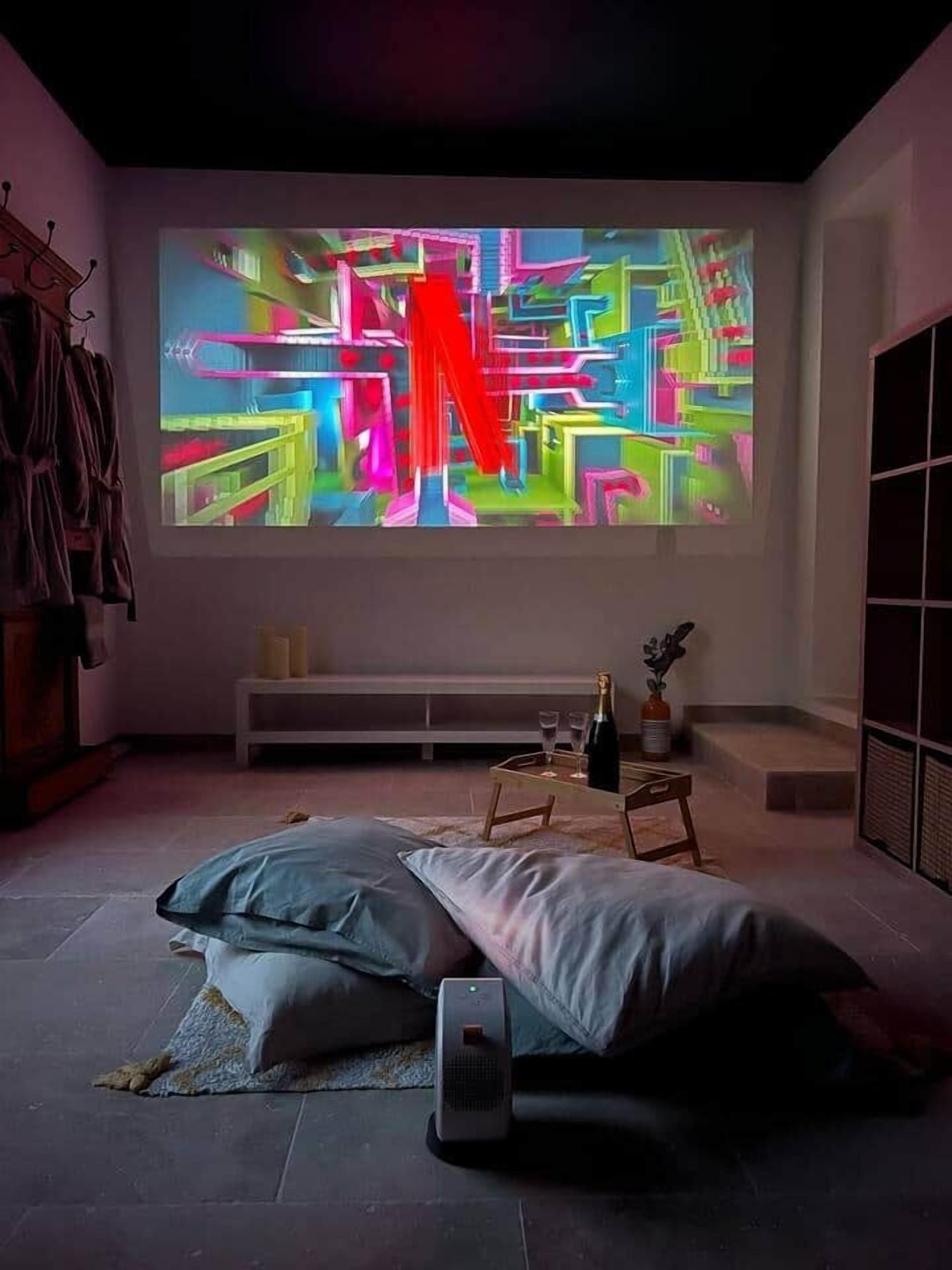 A modern room with a large projection screen displaying colorful abstract art, a bed with pillows, a bedside table with a bottle and glasses, and a small speaker.