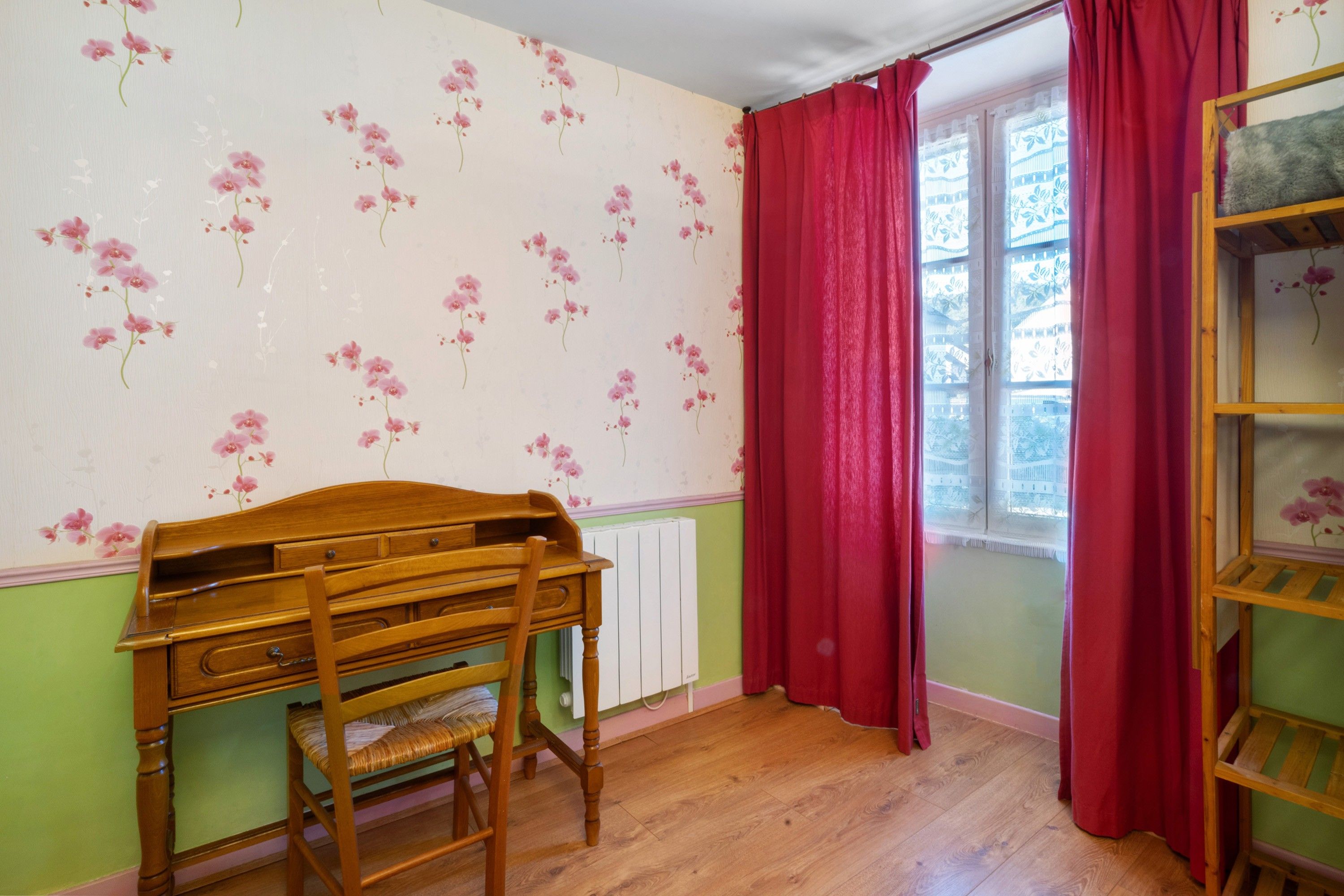 A room with a wooden desk, a radiator, a window with red curtains, and a bookshelf.