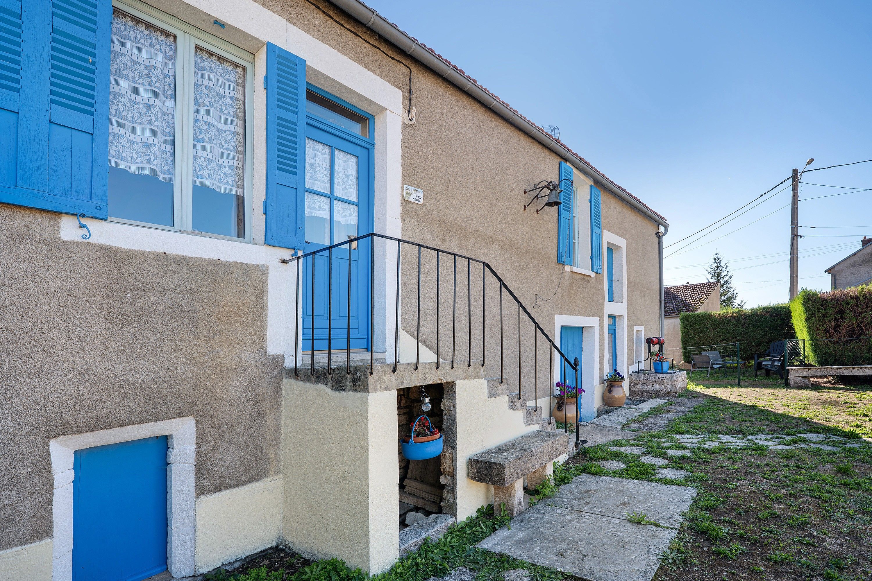 A two-story building with blue shutters, a stone staircase, and a small garden area with a bench.