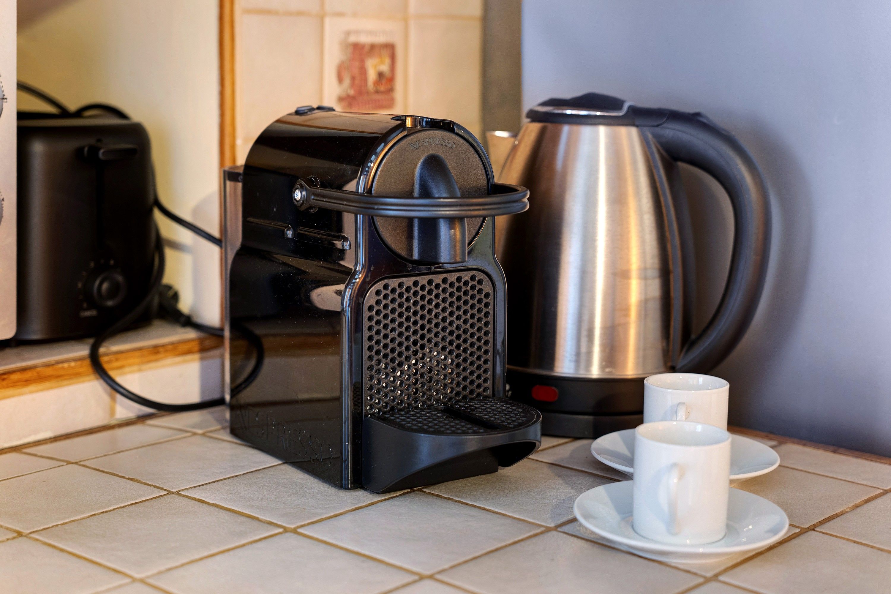 A coffee maker, a kettle, and two white cups on a tiled kitchen counter.