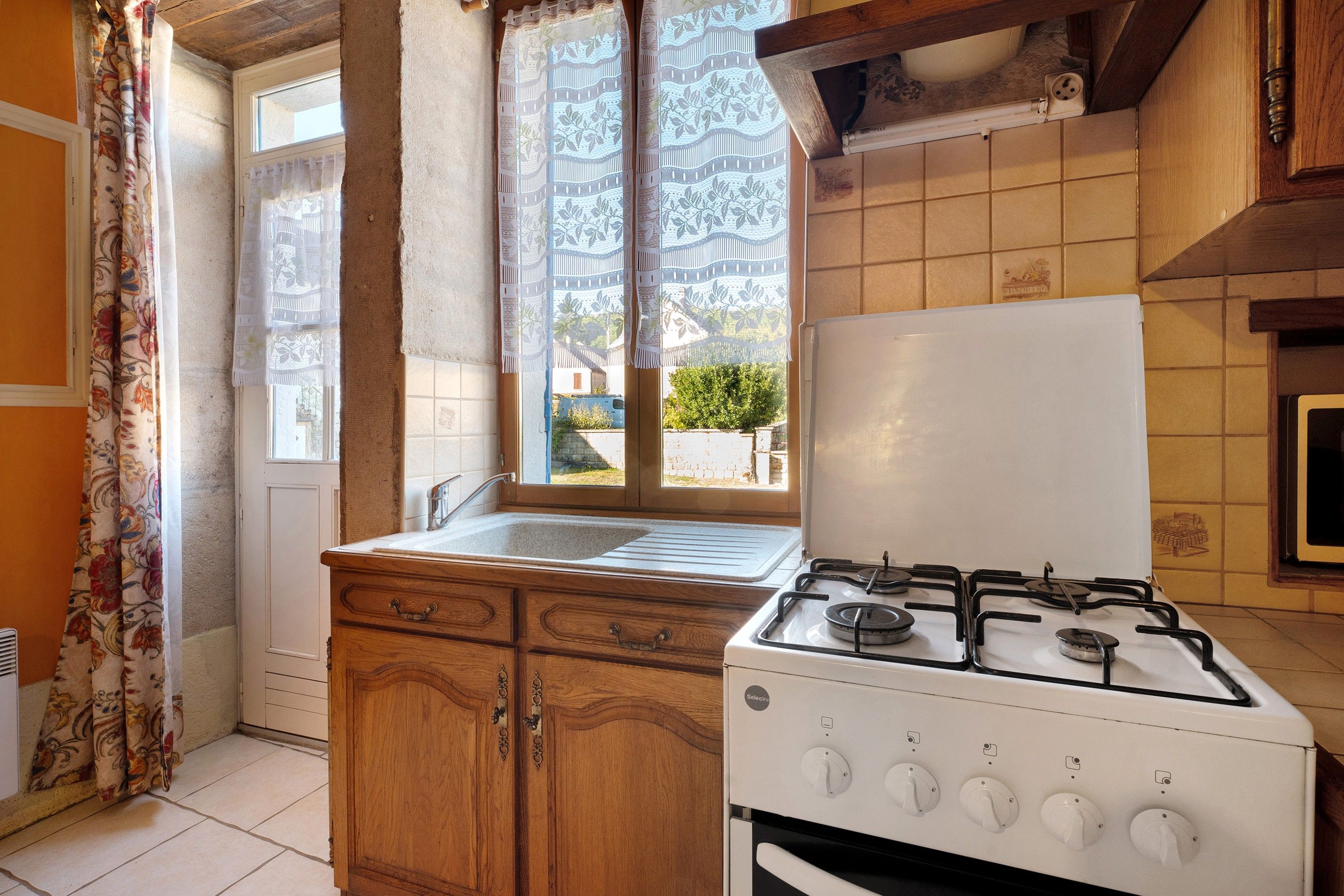 Une cuisine équipée d’une cuisinière à gaz, de meubles en bois, d’une fenêtre agrémentée de rideaux à motifs et offrant une vue sur une maison à l’extérieur.
