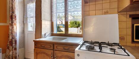 A kitchen with a gas stove, wooden cabinets, a window with patterned curtains, and a view of a house outside.
