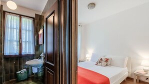 Hotelkamer met een bed, nachtkastje, wastafel, raam met gordijnen en een groene mand.
