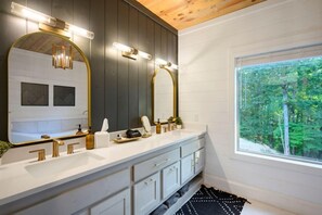 Un baño con tocador blanco, detalles dorados, un espejo grande y una ventana con vistas al bosque.