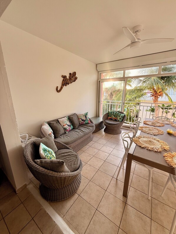 A sunlit room with a sofa, two wicker chairs, a dining table with placemats, a ceiling fan, and a view of palm trees through large windows. - 3-Bedroom Apartment by the Sea in Saint François (Saint-François)