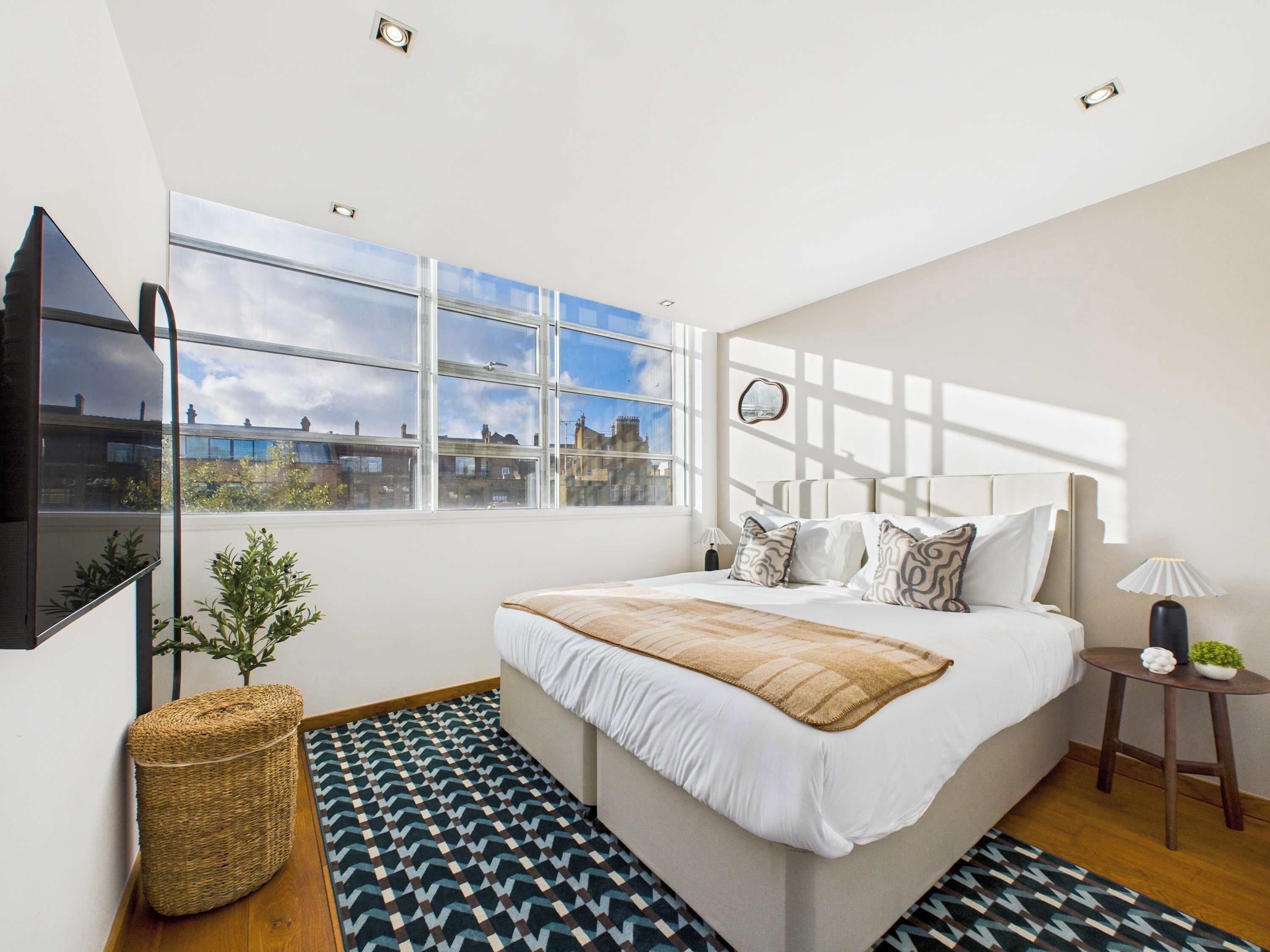 A modern bedroom with a large bed, a TV mounted on the wall, a window with a city view, and a small side table with a lamp.