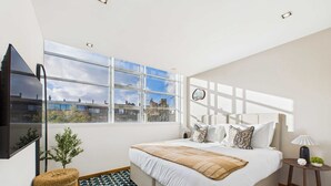 A modern bedroom with a large bed, a TV mounted on the wall, a window with a city view, and a small side table with a lamp. - Light-filled Camden Town Balcony Apartment (Greater London)