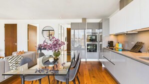 A modern kitchen with built-in appliances, a dining area with a glass table, and a view of the city through large windows. - Light-filled Camden Town Balcony Apartment (Greater London)