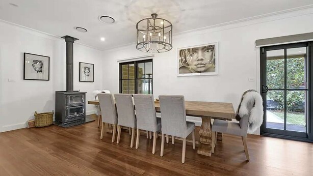 Un comedor con una mesa de madera, sillas tapizadas en gris, un estufa de leña negra y un ventanal grande.