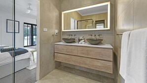 Un baño moderno con un espejo grande, dos lavamanos, una ducha y una puerta de cristal que da a un dormitorio.