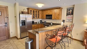 A kitchen with wooden cabinets, a stainless steel refrigerator, a microwave, and a bar area with metal chairs. - Mountaineer Square 313 | 1BR Walk-to-Lifts Condo, Sleeps 4, Pool & Hot Tub (Crested Butte)