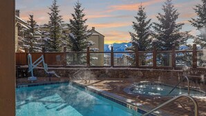 A pool area with hot tubs, a wooden deck, and a view of snow-covered trees and mountains. - Mountaineer Square 313 | 1BR Walk-to-Lifts Condo, Sleeps 4, Pool & Hot Tub (Crested Butte)