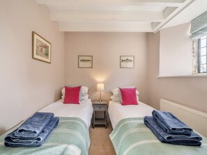 A hotel room with two beds, each with a pillow and a rolled towel. There are framed pictures on the wall, a nightstand with a lamp, and a window with a view.