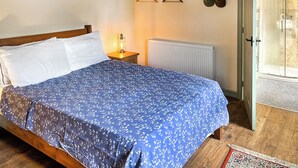 Quarto com uma cama, mesa de cabeceira com abajur, radiador, cabideiro com chapéus e porta que dá para um banheiro.