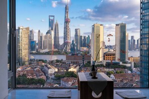 A cityscape view from a high-rise hotel room with a large window, a vase with flowers, and a wooden side table. - Book and Bed - North Bund (Shanghai)