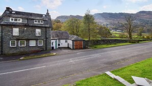 Una casa de piedra con puerta verde y garaje, ubicada en zona rural con carretera y montañas al fondo.