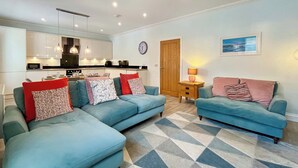 Een moderne woonkamer met twee blauwe banken, een vloerkleed met een geometrisch patroon, een houten deur en een keuken op de achtergrond.