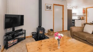 Ein Wohnzimmer mit Fernseher, einem schwarzen Kaminofen, einem Holztisch, einer Couch und einer Küche im Hintergrund.