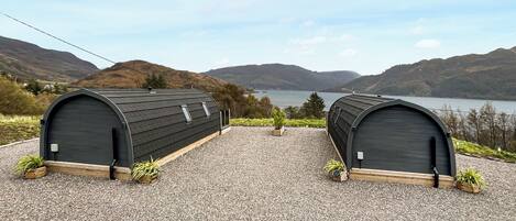 Two small, dark-colored, arched structures with a gravel ground, set against a backdrop of hills and a lake.