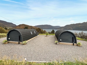 Two small, dark-colored, arched structures with a gravel ground, set against a backdrop of hills and a lake.