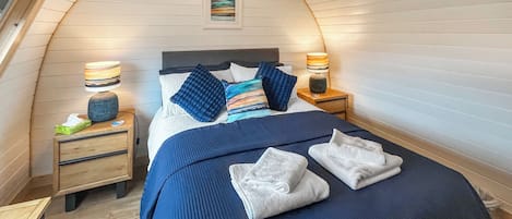 Un dormitorio con techo curvo, una cama con ropa de cama azul, dos mesitas de noche con lámparas y un pequeño cuadro en la pared.