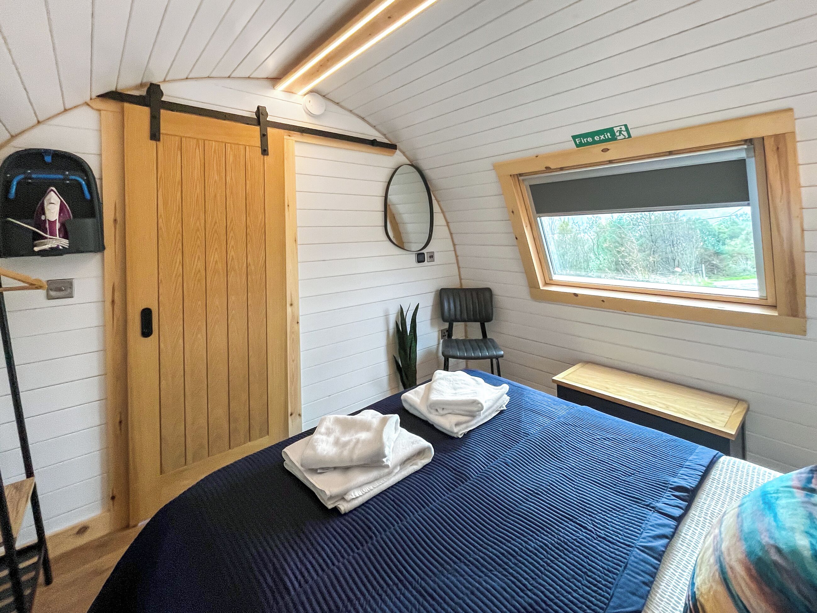 A compact room with a bed, a chair, a small table, a wardrobe, a mirror, and a window with a view of trees.
